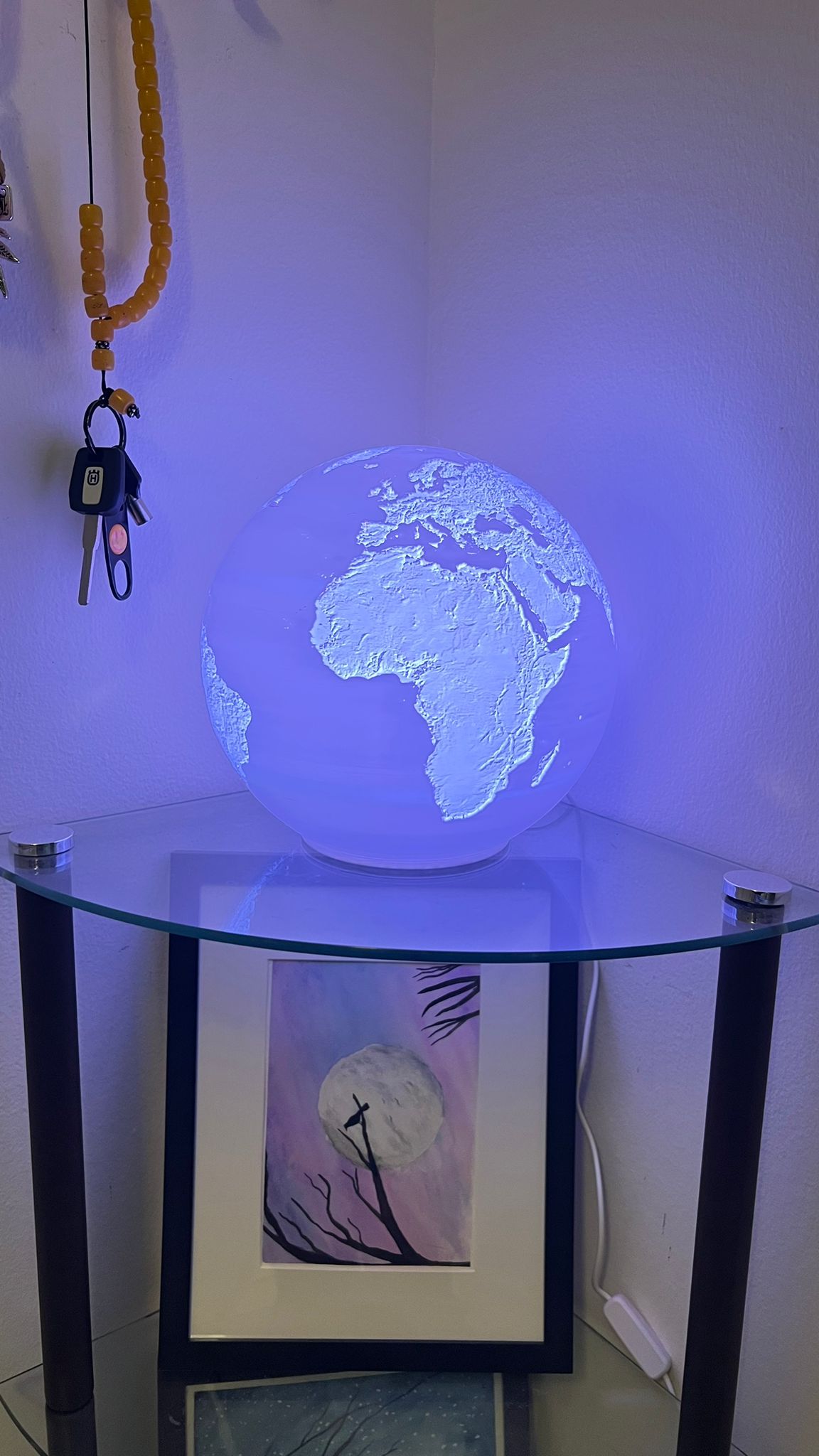 A 3D-printed globe lamp with detailed topography of Earth's continents, illuminated with a soft white glow on a glass shelf