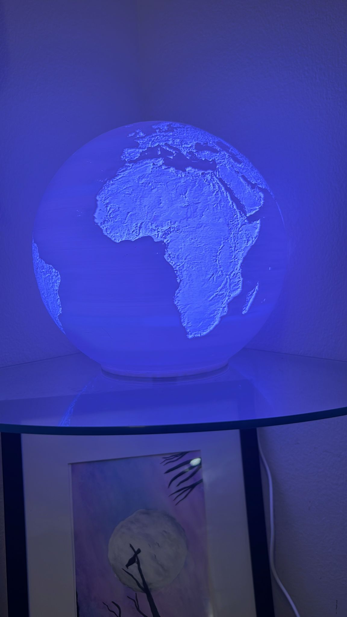 A glowing 3D globe lamp highlighting Earth's continents with a striking blue LED light, placed on a glass shelf in a dimly lit room.